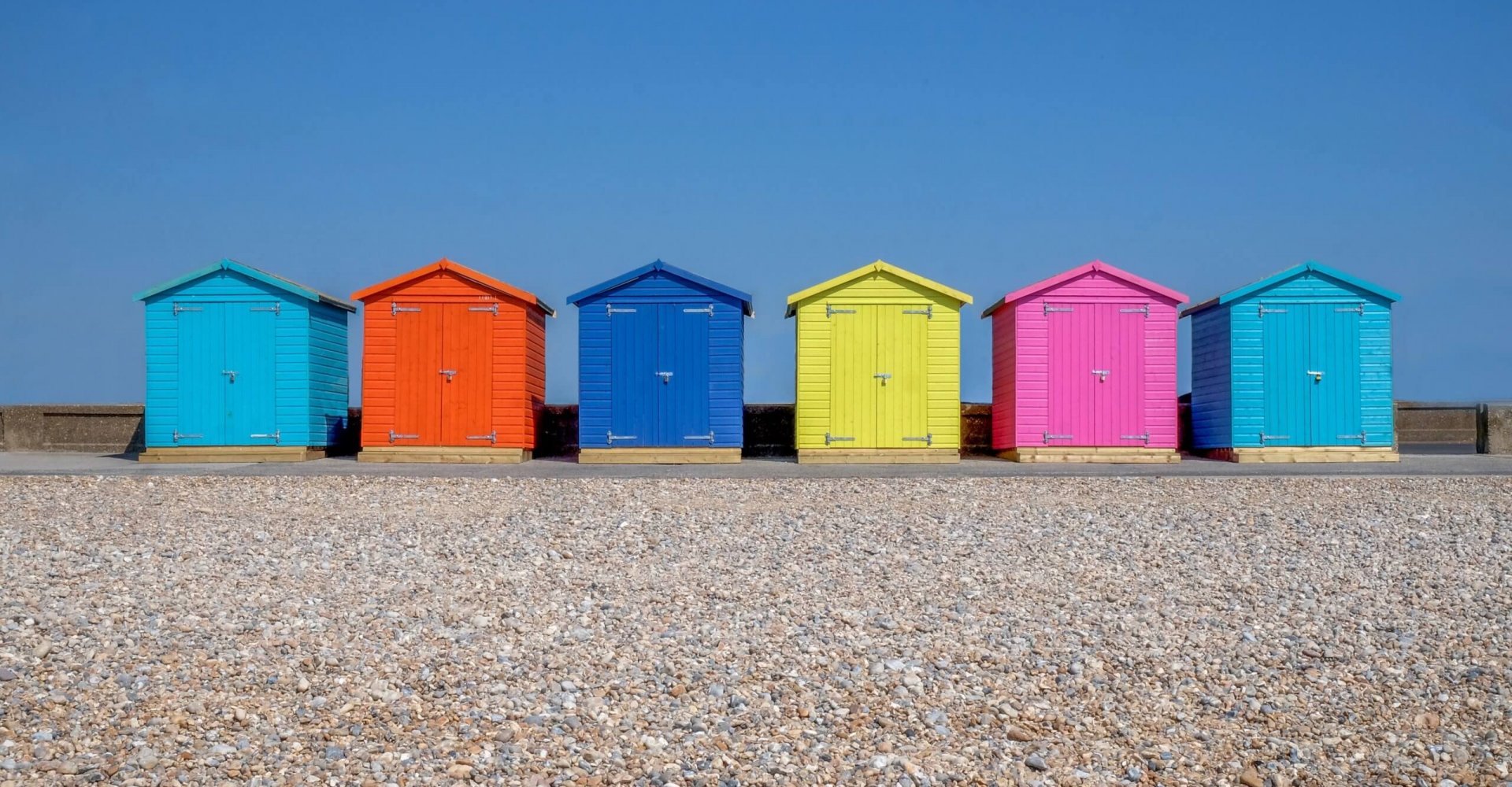 A line of six brightly coloured Beach huts, each one is a different colour in the foreground is a pebble beach and behind is a bright blue sky, Seaford, east Sussex, England, United Kingdom, UK,